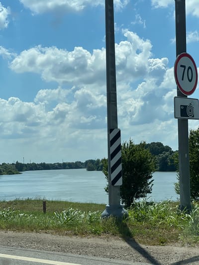 The image is a landscape-oriented photograph taken from a vehicle traveling alongside a body of water, likely a canal or river, in Belozyorny, Russia. The sky is a bright blue with large, fluffy white cumulus clouds. On the left side of the image, a metal pole supports two signs. The top sign is circular with a red border and the number "70" in black, indicating a speed limit. Below it is a rectangular sign with a black border and a graphic depicting a camera with a circular lens and a rectangle, signifying a speed camera zone. Further down the pole, another sign with black and white diagonal stripes warns of a hazard or obstruction.
The body of water occupies the majority of the right side of the frame and appears calm, reflecting the sky. A dense line of green trees forms the far bank of the water, with some structures, possibly buildings, visible through the foliage. The foreground features a grassy verge transitioning into a gravel road with a white line marking the edge. A small, dark signpost with some text is planted in the grass near the water's edge. The lighting suggests it is daytime, with the sun casting shadows, and the weather appears to be clear and pleasant. The overall impression is of a scenic drive through a rural or semi-rural area in Russia.
