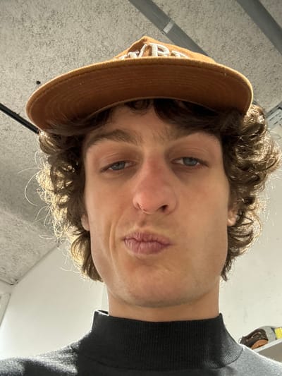 A person's face dominates the frame, taken from a low angle. The individual has light skin, blue eyes, and brown, curly hair. A brown baseball cap is partially visible on their head, with white lettering. Their lips are pursed slightly. They are wearing a dark-colored, high-necked sweater. The background consists of a ceiling with some visible fixtures and a wall. The image's perspective gives a slightly distorted view of the subject's features.
