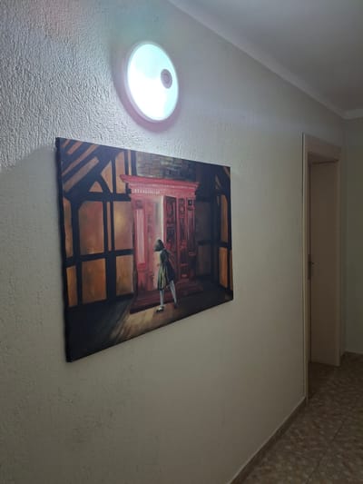 An eye-level, slightly low-angle, indoor shot shows a painting on a wall near a ceiling fixture, possibly in an apartment in Krumovgrad, Bulgaria. The wall is textured and painted light beige. The round ceiling fixture has a white plastic diffuser. Above the painting is a recessed closet or storage area, the bottom of which is flush with the wall. The painting is a rectangular canvas depicting an indoor scene, possibly an interior with a person. The painting's style is somewhat impressionistic, with warm colors dominating the composition. The perspective is slightly skewed, as if the photo was taken at an angle to the wall. The ceiling on the upper-right of the frame transitions into a wall with light beige tiles.
