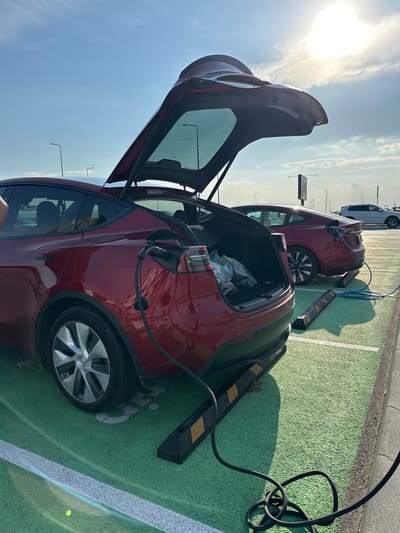 This forensic description details a daytime scene at a charging station in Kapitan Andreevo, Bulgaria. The image captures two red Tesla vehicles, both actively charging in designated green-painted parking spaces, under a bright sunlit sky with scattered clouds.
In the foreground, a dark red Tesla Model Y, recognizable by its open rear hatch and crossover body style, is parked with its rear facing the viewer. The trunk is open, revealing a plastic bag and a water bottle inside. A black charging cable is plugged into the vehicle's charging port, located on its rear left side, and extends down to the ground. Directly beneath the rear bumper, yellow and black striped wheel stops delineate the parking spot. The vehicle's silver multi-spoke wheel is visible on the left.
Behind the Model Y, a second red Tesla, a Model 3 sedan, is also parked and charging. A coiled blue charging cable is connected to its rear left charging port. Similar yellow and black striped wheel stops are present beneath its rear bumper.
The parking area features a distinctive light green, textured asphalt surface, marked with white lines defining the parking bays. Long shadows, indicative of a low sun angle (likely morning or late afternoon), are cast across the ground towards the viewer. In the mid-ground to the right, a white SUV is parked further away.
The background reveals a clear blue sky with some wispy white clouds. The sun, a bright luminous disc, is prominently visible in the upper right, contributing to the strong light and distinct shadows. Several dark, slender utility poles or streetlights are visible against the sky, along with a larger, dark rectangular sign structure in the distance on the right. No individuals are visible in the scene.