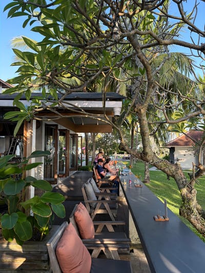 An outdoor seating area at a cafe or restaurant is depicted, with a long, dark grey counter stretching across the foreground. Several patrons are seated at the counter, engaged in conversation or enjoying drinks. The cafe's interior is visible behind the counter, featuring large glass doors and wooden accents. Wooden benches and tables are arranged on a raised wooden deck. Lush greenery, including palm trees and large-leafed plants, surrounds the area, suggesting a tropical environment. In the background, a small building with a terracotta roof is visible, along with a swimming pool. The lighting indicates it is daytime, with sunlight casting shadows and illuminating parts of the scene. The location is identified as Pekutatan, Indonesia.