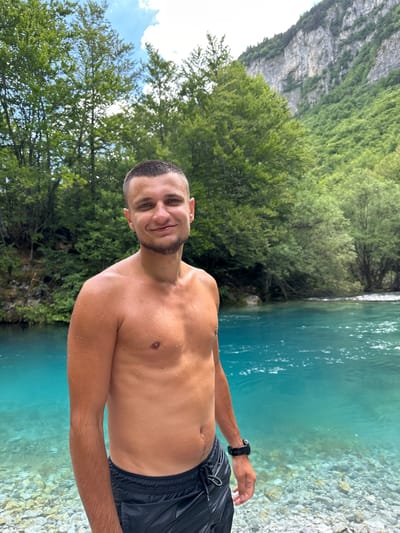 A young man, bare-chested and with short hair and a beard, stands in the foreground of a vibrant turquoise river in Vusanje, Montenegro. Water droplets are visible on his skin, suggesting he has recently been in the water. He is wearing black swim shorts and a black watch on his left wrist. He is smiling at the camera. Behind him, the river flows, its clear water revealing a rocky bed. Lush green trees line the riverbank, and in the background, a majestic, rocky mountain face rises up, partially covered in vegetation, under a cloudy sky.