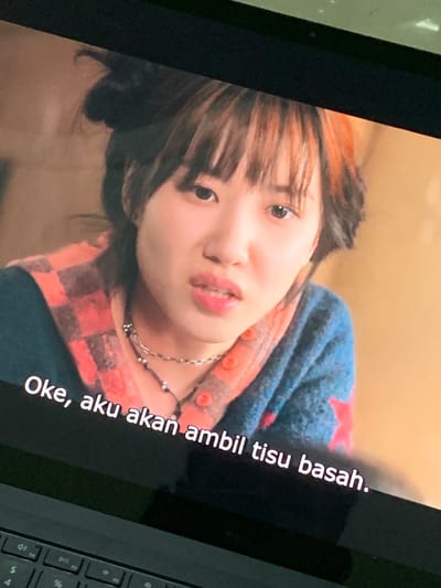 A close-up, head-and-shoulders shot shows a young East Asian woman with bangs and dark hair streaked with reddish-orange highlights. She is wearing a blue and red plaid shirt over a blue top and a necklace. She is looking directly at the camera with a slight grimace and has bright pink lipstick on. White subtitles at the bottom of the screen read "Oke, aku akan ambil tisu basah." The background is a warm, muted orange. The image appears to be a screenshot from a video being displayed on a dark-colored laptop with a visible keyboard in the foreground. The presence of subtitles suggests the media is being watched in the city of Parung Panjang, Indonesia, where Indonesian is spoken.