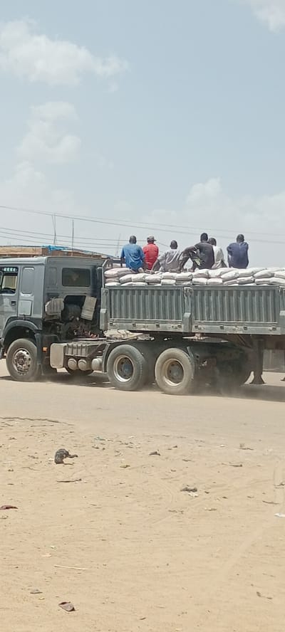 A gray, flatbed truck is transporting a load of what appears to be bags of cement. Six people are sitting atop the bags. The truck is driving on a dirt road in what appears to be a built-up area, with utility lines in the background. There are several pieces of litter on the road in the foreground. The sky is blue with scattered clouds. The location is Gayawa, Nigeria.