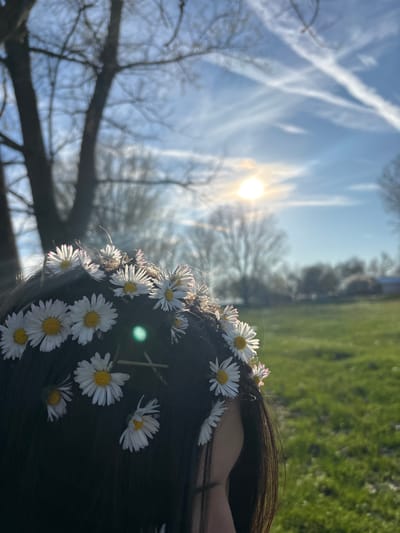 The image, presumably taken in Beli Brod, Bulgaria, focuses on the top of a person's head adorned with a garland of daisies. The person has dark hair. The background is blurred, but it appears to be an outdoor scene with a sky showing sun and contrails, bare trees, and greenery suggesting a park or field.