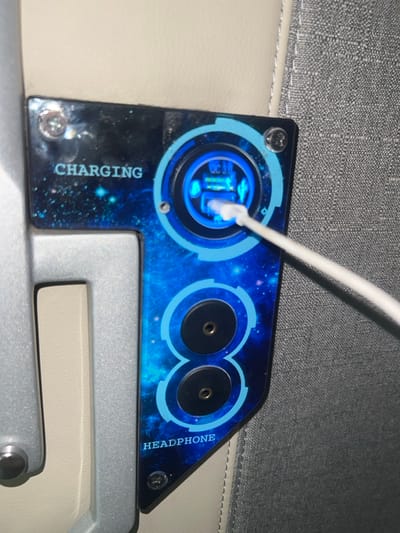 The image displays an integrated multi-function panel within an interior environment, located in Aê Thi, Vietnam. The panel features a dark blue surface with a starfield pattern, secured by four visible screws.
The upper portion of the panel is labeled "CHARGING" in white text. Within this section, a circular module emits a bright blue light, indicating active function. This module contains two stacked USB-A ports, one of which has a white USB-A cable inserted, glowing with blue light at the connection point. The visible label "QC 3.0" indicates Quick Charge 3.0 capability for the USB ports.
The lower portion of the panel is labeled "HEADPHONE" in white text and contains two vertically arranged 3.5mm audio jacks. These jacks are black with visible gold-colored internal contacts.
The panel is flanked by structural and upholstered elements. To its left, a light grey, textured frame or support structure is visible, behind which a segment of cream-colored upholstery is present. To its right, a grey, tightly woven fabric, consistent with seat upholstery, is visible. No individuals are depicted in the scene.