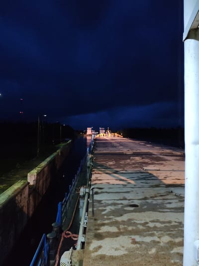 A nighttime shot depicts the exterior of a lock, likely part of a canal system, in the village of Devyatiny, Russia. The scene is illuminated by artificial lights, casting shadows and highlighting the wet, industrial surface of what appears to be a barge or lock gate. The water within the lock is dark and still. In the distance, illuminated structures suggest operational equipment, possibly part of the lock's machinery or a nearby building. The sky is a deep, dark blue, indicating night. The environment is characterized by concrete structures, metal railings, and bollards, consistent with a functional waterway. No individuals are clearly visible in the frame.