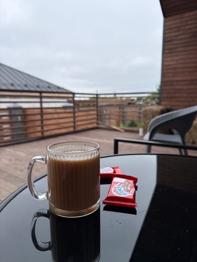 A glass mug filled with coffee sits on a black reflective table. Two red candy wrappers are next to the mug. The table is on a wooden deck, which appears to be part of a porch or patio. In the background, a wooden railing separates the deck from a sloped roof structure. Beyond the railing, there's a cloudy sky and some greenery. The scene is likely outdoors, on the porch of a house or cottage, possibly in the city of Semiluki, Russia, given the architectural style and natural surroundings.