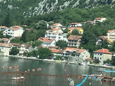 The image shows a waterfront scene in Donji Orahovac, Montenegro. The foreground features a body of water with several orange buoys and some debris in the water, possibly part of a dock or barrier. A rusty metal structure with vertical supports and horizontal bars runs across the lower portion of the image.

Behind the water, a shoreline is visible with numerous buildings, many of which appear to be houses or residential buildings. These structures are nestled into the side of a steep, tree-covered hill. The architecture varies, with some buildings featuring red tile roofs and others white or light-colored facades. Some buildings have multiple stories, and balconies or terraces are visible on some.

Trees and dense green foliage cover the hillside behind the buildings, creating a lush backdrop. The vegetation appears to extend up the rocky slopes of the mountain. Several boats are docked or moored along the shoreline, including at least one white motorboat. A few people are visible on the shore and near the water, some appearing to be enjoying leisure activities. The overall impression is of a Mediterranean coastal town or village situated at the base of a mountainous terrain.