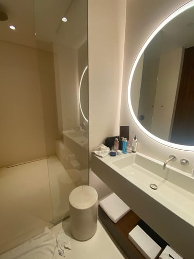 The image depicts a bathroom interior, likely in a hotel or residential setting. There's a brightly lit, round mirror with an illuminated border. Beneath the mirror is a modern, white sink with a stainless steel faucet. A small shelf to the side of the sink holds toiletries.
To the left is a shower enclosed by a glass partition. A white, cylindrical stool sits in front of the glass partition. A white towel is on the floor near the stool. The location of the bathroom is El Caracol, Mexico.