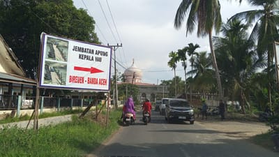 The image depicts a street scene in Blang Mee, Indonesia, dominated by lush tropical vegetation, particularly palm trees. A prominent sign on the left advertises "JEMBATAN APUNG KEPALA HIU" (Floating Bridge Head Shark) with directional arrows pointing right and indicating "BIREUEN - BANDA ACEH." The sign also features two images of a floating bridge.
In the foreground, a paved road runs through the scene. Two motorcycles are visible on the road. The first, closer to the viewer, is ridden by a person wearing a purple headscarf and light-colored clothing. The second motorcycle, slightly ahead, is ridden by a person in a red shirt. A black car follows them. In the background, a distinctive pink domed mosque stands out against the cloudy sky, with other buildings and more palm trees surrounding it.
The environment appears to be a rural or semi-urban area, characterized by the presence of a road, buildings, vegetation, and electrical poles and wires. The overall atmosphere suggests a bright, possibly warm day, given the clear visibility.