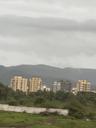 The image captures a cloudy, overcast day in Palghar, India, with a landscape featuring rolling hills in the background. In the midground, several apartment buildings are visible, varying in architectural style and color. The buildings are situated amidst lush green trees and vegetation. In the foreground, there is a grassy area with a low, white concrete wall running horizontally across the scene. The overall impression is one of a developing urban area nestled within a natural, hilly environment.
