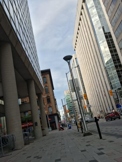 This image depicts an urban scene, likely in Ottawa, Canada, with a focus on a city street and surrounding buildings. The perspective is angled, with a tall, modern building featuring a glass facade dominating the left side of the frame. Its reflective surface mirrors the sky and other structures.
A street runs vertically through the image, lined with a patterned sidewalk. Several cars are visible on the street, including a red vehicle and a silver car. Traffic lights and street signs are present, indicating an active intersection. The architecture of the buildings varies, with some appearing more contemporary and others older, constructed from brick.
The time of day appears to be either early morning or late afternoon, judging by the quality of light. The sky is partly cloudy. There are no people clearly visible in the image, suggesting the activity is primarily related to traffic and the urban environment itself.
Notable details include the grid-like pattern of the sidewalk, the sleek lines of the modern building, and the reflections in its windows. The presence of bicycle racks hints at a mode of transportation common in urban settings. Text is visible on a sign in the background, appearing to be in red lettering. The overall impression is one of a bustling city street, captured from a dynamic, slightly disorienting angle.