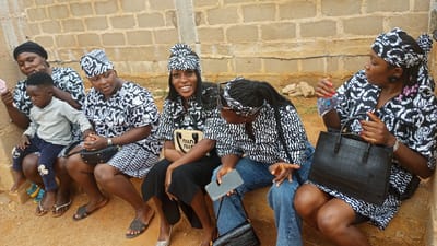 Five women and one child are sitting together outside. The women are wearing matching black and white patterned dresses and head coverings. The child is wearing a gray sweatshirt and dark blue pants. They are sitting on a dirt-colored ledge in front of a cinderblock wall. Some of the women are holding handbags or phones. The location is Barikin Sabon Gidan Kanat, Nigeria.