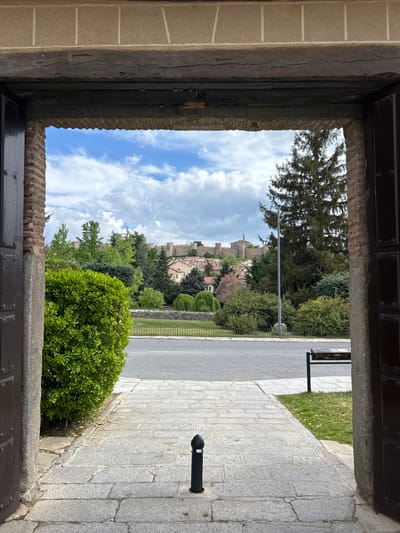 View of the medieval city wall of Ávila, Spain, through an arched doorway. The foreground features weathered stone and wood framing the doorway. Outside, a street runs parallel to the wall. Green trees and bushes line the street. The sky is blue with scattered clouds. A bollard stands on the stone pavement.