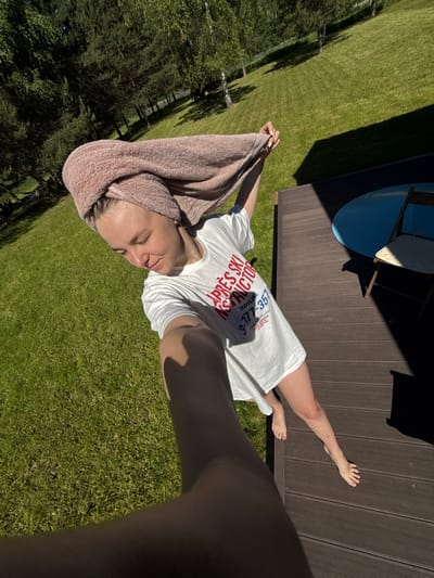 The image captures a young woman standing on a wooden deck outdoors in Копнино, Russia, under bright sunlight. She is taking a selfie, holding the camera in her outstretched right hand, which is prominently featured in the lower left foreground. Her head is wrapped in a light pink terry cloth towel. She is wearing a loose white t-shirt with red lettering spelling "APRES SKI INSTRUCTOR" and blue text that includes "HOTLINE 3-177-357". Her bare legs and feet are visible, with her left foot resting on the deck and her right foot slightly lifted. Her gaze is directed downwards, and her expression is tranquil.
The dark brown wooden deck occupies the right and lower sections of the frame. A deep, angular shadow from an unseen structure extends across the deck, obscuring a blue circular table and a wooden chair with a light cushion positioned in the upper right. Beyond the deck, a lush, green grassy lawn slopes gently upwards into the background. Several trees, including light-barked birches and darker conifers, are scattered across the distant landscape, with shadows cast upon the grass. The scene is brightly illuminated, with clear distinctions between sunlit areas and sharp, dark shadows.