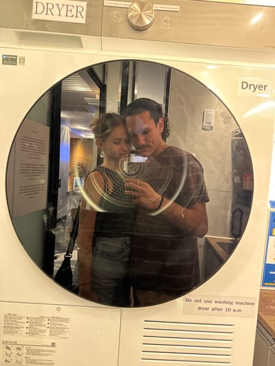The image depicts a reflection of two individuals captured in the glass door of a white, front-loading Samsung dryer, confirming its location in South Korea due to visible Korean text on labels. Given the instruction, the setting is identified as Jongno-gu, South Korea, likely within a shared accommodation's laundry facility.
The dryer features "DRYER" labels, a control knob, and buttons with "ON/OFF (Press for 3 seconds)" and "PLAY (Press for 1 second)" instructions. A prominent warning sticker below the door states, "Do not use washing machine dryer after 10 p.m."
Reflected in the dryer door are a light-skinned female and a light-skinned male. The female, positioned on the left, has light brown hair, wears a dark top and light denim shorts, and has visible tattoos on her left arm. She is holding a smartphone with both hands, angled as if capturing the reflection, indicating a selfie. A black strap, possibly from a bag, is visible over her right shoulder (viewer's left). The male, positioned on the right, has dark, wavy hair, a mustache, and is wearing a striped short-sleeved shirt. His left arm is around the female, with his hand visible near her left shoulder. Both individuals are looking down towards the smartphone, their faces close together, indicating an intimate interaction.
The background reflected in the dryer door reveals an indoor environment. On the left, behind the female, a wall with partially legible text and a black object (possibly a bag or strap) is visible. Further back, wooden paneling, artificial lighting, and potted plants suggest a common area. On the right, behind the male, a light switch and a countertop area are discernible.