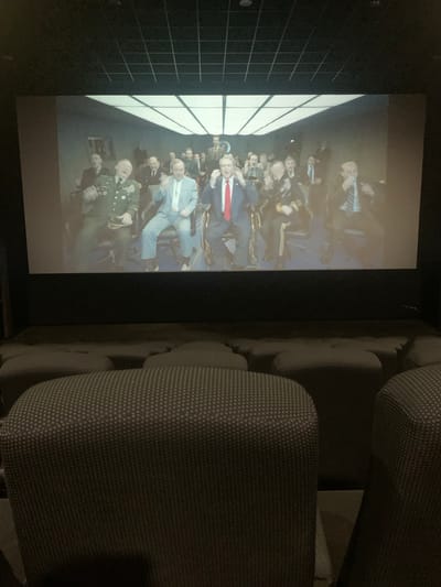 In a dimly lit, modern movie theater, the large screen displays a scene of men in formal attire, some in military uniforms, seated and appearing engaged. The foreground is dominated by rows of dark, upholstered theater seats, some appearing to be recliners. The environment suggests a viewing of a film or presentation. There is no indication of the location being in Sinalunga, Italy.