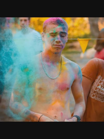 The image depicts a young man covered in colorful powders, likely during a Holi festival or a similar celebration. He is shirtless, revealing his chest and arms, which are smudged with various hues of pink, blue, green, and yellow. His eyes are closed, and his head is tilted slightly upwards, suggesting a moment of sensory experience or contemplation. His hair is also tinged with pink powder. He is wearing a silver chain around his neck and a black bracelet on his left wrist.

To his left, a cloud of blue and teal powder billows around him, obscuring part of his body and the background. In the background, other individuals are visible, though they are out of focus. One person in the middle ground is wearing a white shirt, and another person to the right is wearing an orange t-shirt with repeating text that appears to read "GLAM." A yellow triangular flag is also visible in the background, adding to the festive atmosphere. The overall setting appears to be outdoors, with a natural, slightly blurred environment suggesting a park or open area.

The image does not provide any specific visual cues or landmarks that would definitively place the scene in the city of Norcia, Italy. Therefore, without additional information, it is not possible to specify the location as Norcia.