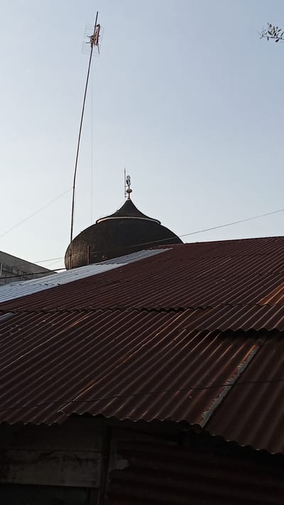 In the city of Geulanggang Teungoh, Indonesia, a scene unfolds featuring the prominent dome of a mosque, partially obscured by two weathered, corrugated metal roofs in the foreground. The larger roof, a rusty red hue, occupies the lower portion of the frame, with its rippled surface catching the light. Behind it, a second, lighter colored corrugated roof juts out from the left. The mosque's dark, conical dome rises above the roofs, topped with a crescent moon and star finial. A tall, thin antenna, also appearing weathered, extends upwards from behind the mosque's dome, its horizontal element reaching towards the pale, clear sky. The sky itself is a soft, washed-out blue, with a faint suggestion of light filtering from the setting or rising sun, creating a warm glow around the edges of the dome. A few distant, indistinct tree branches are visible in the upper right corner, adding a touch of natural element to the otherwise man-made landscape. The overall impression is one of a typical urban or semi-urban scene in Indonesia, where religious structures and everyday building materials coexist.
