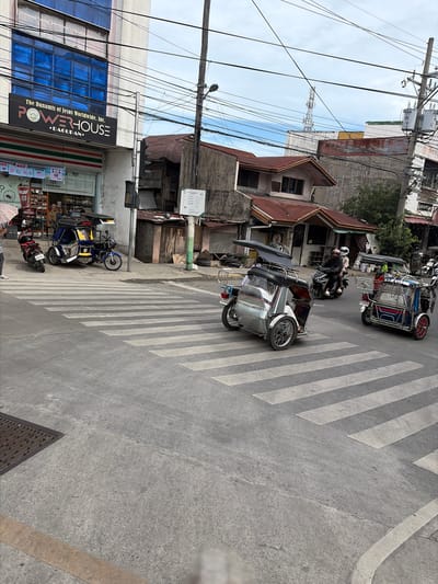 The image depicts a street scene in Dagupan City, Philippines, likely a commercial area, on an overcast day. A pedestrian crossing is prominently featured in the foreground, with several motorized tricycles navigating the intersection.
On the left side of the image, a multi-story building with blue tinted windows is visible, housing a 7-Eleven convenience store on the ground floor and a sign that reads "The Dunamis of Jesus Worldwide, Inc. POWERHOUSE • DAGUPAN".
Several tricycles, a common mode of transport in the Philippines, are present. One is blue and yellow, parked near the sidewalk. Another, a silver tricycle, is in motion, crossing the pedestrian lane. A third tricycle, predominantly blue with red accents, is also crossing the pedestrian lane. Two individuals are visible riding a motorcycle and another tricycle, all appearing to be in transit.
The environment is urban, characterized by concrete roads, sidewalks, buildings, and overhead electrical wires. The presence of a 7-Eleven and signs in English suggests a developed commercial area. The overall impression is of a typical day in a bustling Philippine city.