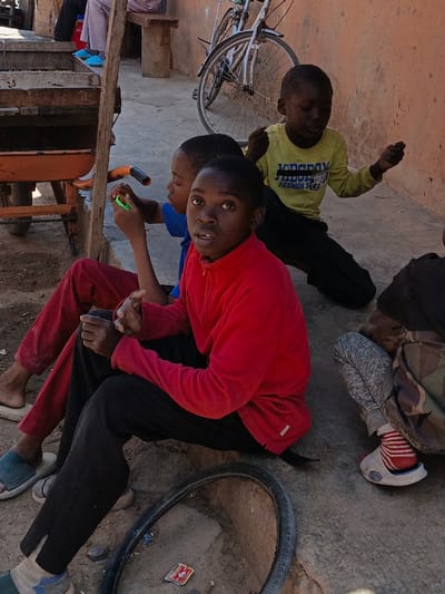 This image depicts an outdoor scene in the city of Gayawa, Nigeria, featuring multiple individuals, primarily children, in varying states of repose on a concrete and dirt ground.

In the foreground and central to the image, a young male, appearing to be a child, is seated, facing forward with a direct gaze toward the observer. He wears a red, long-sleeved top with a quarter-zip collar and black trousers. To his left, another young male, also a child, is seated, wearing a blue short-sleeved top and red trousers. He holds a bright green, cylindrical object in his right hand. Directly behind this second child, a wooden structure, possibly part of a cart or stand, is visible, featuring an orange handle. Further to the right, a third young male child is seated, dressed in a yellow long-sleeved shirt with "JADEDAY FASHION S" text and black trousers; he holds thin, light-colored strings in both hands. The lower body of a fourth individual, partially obscured on the far right, shows camouflage-patterned trousers and feet clad in red and white striped socks within white sandals.

In the immediate foreground, a detached black bicycle tire rests on the ground, near a small, red, rectangular object, possibly a wrapper or matchbox. The ground surface transitions from rough concrete to packed dirt. In the background, leaning against a light orange-brown wall, a bicycle with a silver frame and black tires is visible. To the far left in the background, a person's lower legs are visible, seated on a wooden bench, wearing light blue sandals.