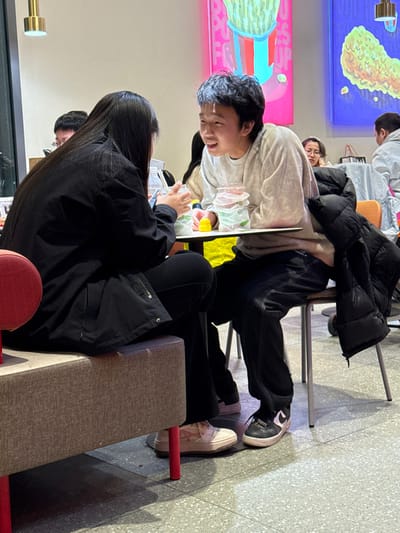 The image displays an indoor scene within a casual dining establishment in Changping District, China. In the foreground, two individuals are seated at a dark-surfaced table. On the left, a person with long, dark hair, wearing a black long-sleeved top and black pants, sits on a light grey upholstered bench. This individual's left hand holds a clear plastic cup, and they are wearing light pink and white athletic shoes. Facing them on the right, a male with short, dark hair, wearing a light grey hooded sweatshirt and dark pants, sits on a metal-frame chair with an orange back. He is smiling broadly with an open mouth, looking towards the individual on the left, indicating active conversation. A black puffer jacket is draped over his chair. On the table in front of him are two stacked clear plastic cups with green and white branding, a small yellow duck-shaped object, and a water bottle. He wears dark athletic shoes. In the background, other patrons are visible seated at additional tables. The walls are light-colored and decorated with two large, brightly colored framed artworks depicting stylized food items, one predominantly pink with green elements, and the other blue with a yellow/green item. The floor is light grey and speckled.