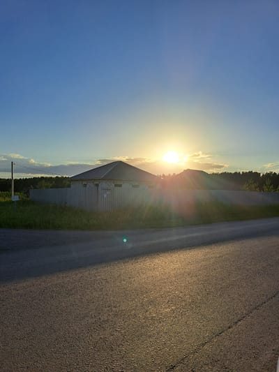 The image depicts a rural or suburban scene under a bright sky, likely in Chernetsk, Russia, with the sun prominently featured. In the mid-ground, a long, grey metal fence encloses a property. Behind the fence, a light-colored building with a dark roof is visible, suggesting it might be under construction or a simple outbuilding. There are trees in the background, forming a dark treeline against the horizon. The foreground is dominated by an asphalt road, reflecting the sunlight and creating a bright, shimmering effect. A single, small green lens flare is visible on the road. The sky is a clear blue, with some wispy clouds near the horizon where the sun is setting or rising. The overall impression is one of a quiet, undeveloped area on the outskirts of a town or city.