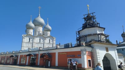 This is a full-shot, eye-level, outdoor, daytime, wide-angle photograph of the Rostov Kremlin in Rostov, Yaroslavl Oblast, Russia. The sky is a clear, bright blue with no clouds. In the background, on the left, is the Cathedral of the Assumption, a white stone structure with multiple silver-domed towers topped with golden crosses. To the right of the cathedral is a white building with a white archway and an ornate black gate. This building appears to be under renovation, with wooden scaffolding surrounding its upper section. In front of this building is a low-lying, orange structure with several arched entrances and awnings. Signs in Russian are visible on the orange building, indicating shops. There are several people in the scene. In the foreground, on the right, a woman with blonde hair is facing away from the camera, looking to the left. Near the center, standing in front of the orange building, are four individuals, two women and two men, facing the building and looking at what appears to be a map or information board. A fifth person is partially visible to the far right. The ground in the foreground is paved.