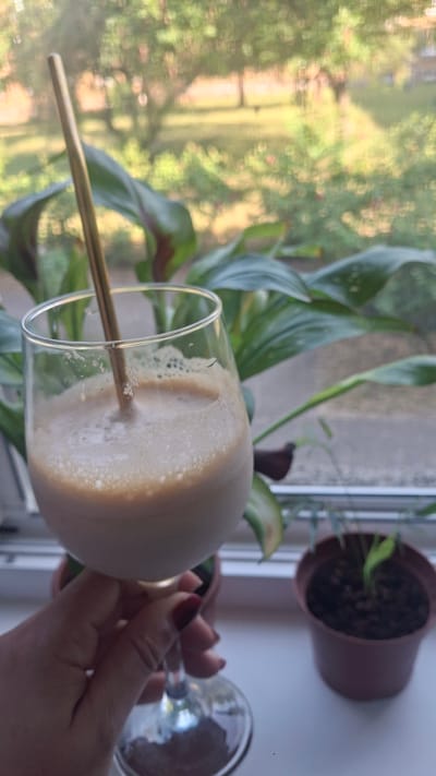 A hand with dark red nail polish holds a stemmed glass containing a frothy, light brown beverage. A golden straw is submerged in the drink. The glass rests on a white windowsill, behind which large, glossy green leaves of a plant are visible. Beyond the plant, through a window with a screen, a blurred outdoor scene of trees and greenery is apparent, bathed in soft daylight. Two small potted plants with dark soil sit to the right of the glass on the windowsill. The overall impression is a casual, indoor setting with a view of nature.