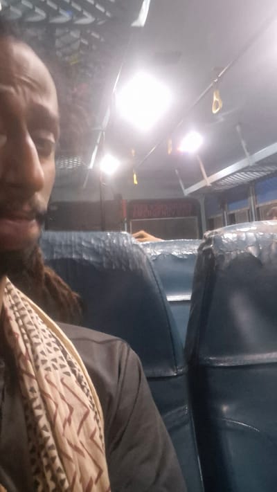 The image shows a person lying down on what appears to be a seat, possibly on public transportation, given the presence of luggage racks and blue upholstered seating. The lighting is bright, suggesting indoor artificial light rather than daylight. The person's face is partially visible and appears to be in repose, perhaps sleeping or resting. They are covered by a patterned fabric. In the background, there are more seats and some signage, though the text is not legible. The overall scene is dimly lit except for the bright overhead lights. The location context is Kullu, India.