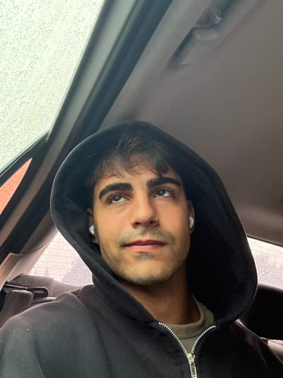 A man wearing a black hooded sweatshirt is seated inside a car, likely in Quilmes Oeste, Argentina. It is raining, as evidenced by the water droplets on the car window. The man has dark hair and is wearing earbuds. The image is taken from a low angle.
