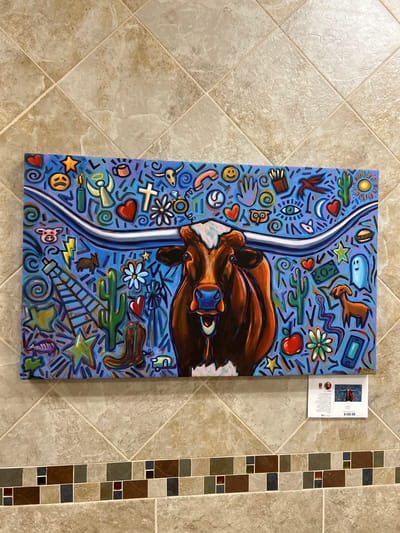 The image shows a colorful acrylic painting of a Texas Longhorn cow with large horns set against a blue background filled with various small icons and symbols. The cow is depicted from the chest up, with its head turned slightly to the viewer's right. Its eyes are wide open, and its mouth is open in a smile, revealing its teeth. The background is a vibrant blue and is adorned with a multitude of colorful, cartoonish illustrations, including a smiling sun, various hearts, stars, lightning bolts, a cowboy boot, cacti, an owl, an eye, a moon, a ghost, a hamburger, a phone, and a dollar bill, among others. The painting is framed by light brown and beige square tiles on the wall, with a decorative border of smaller, multi-colored square tiles below the painting. A small white card with text and an image of the painting is attached to the bottom right corner of the artwork, displaying a price of "$189.98". The painting is hung on a wall in what appears to be an interior space, possibly an art gallery or a shop.