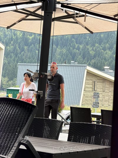 This image, taken in Trnovo Municipality, Bosnia and Herzegovina, captures an outdoor scene at what appears to be a cafe or restaurant. The photo is oriented vertically, with the landscape and a building in the background, framed by outdoor seating and overhead structures.
Two people are the primary subjects. A man, wearing a grey t-shirt and dark shorts, is positioned higher in the frame, looking towards the camera with a slight smile. He is wearing a red baseball cap. A woman, in a white t-shirt and dark pants, stands below him, also looking down and slightly away. She has dark hair and wears glasses. She carries a coral-colored bag over her shoulder. They appear to be patrons or perhaps staff at the establishment.
The setting includes a covered outdoor area with a beige retractable awning supported by a dark metal frame. Black wicker chairs and tables are visible in the foreground and mid-ground, suggesting a dining or seating area. Behind the seating, a building with a wooden facade and grey siding is visible, with a sloping roofline. The background is dominated by a dense, green forest, indicating a mountainous or hilly region. A large green utility box with some signage is also present.
The lighting suggests it is daytime, possibly late morning or early afternoon, with diffused light, and the presence of trees and the overall greenery hint at a pleasant, possibly temperate weather. The scene feels relaxed and casual.
Visible text is limited to small labels and signs on the building and potentially the utility box, but they are too small and indistinct to be read. The overall impression is of a serene, natural setting combined with an inviting outdoor seating area.
