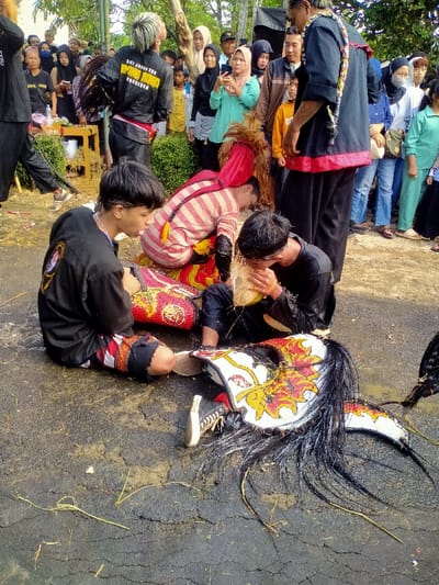 Three people are on their knees on the ground, surrounding a traditional costume. One of the people is drinking something from a coconut, while the others appear to be in a trance-like state. Spectators surround the scene, with some filming the spectacle with their phones. The event seems to be taking place in an outdoor setting, as green plants and trees are visible in the background.