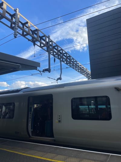 An exterior, low-angle, full shot captures a modern train at the Luton train station platform, under a bright blue sky with scattered white clouds. The train, a sleek, silver passenger carriage, has its doors open, revealing passengers boarding with luggage. The train's windows reflect the sky and the surrounding structures.
Above the train, overhead power lines and a complex metal support structure dominate the upper portion of the frame, indicative of an electrified railway. To the right, a contemporary building with dark, horizontally-paneled cladding rises, contrasting with the open sky and the industrial overhead infrastructure. The platform is visible in the foreground, with yellow and white tactile paving lines marking the edge. The scene appears to be captured on a clear day.