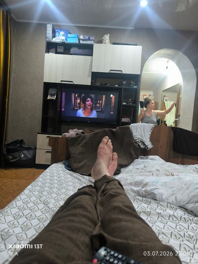 The image depicts a bedroom scene, likely in a residential setting in Orlovka, Russia. The viewpoint is from a person lying in bed, looking towards a television screen mounted on a wall unit. In the foreground, the legs and feet of the person are visible, clad in brown pants, resting on a bed with patterned white sheets.

On the wall unit, a television displays a woman speaking, suggesting a movie or show is in progress. Various items are placed on the shelves of the wall unit, including boxes, decorative objects, and what appears to be a framed picture. To the left of the television, a dark grey duffel bag rests on the floor.

In the background, an arched doorway leads to another room where a woman is standing in front of a mirror, reaching out towards it. She is wearing a grey tank top. The overall environment suggests a domestic living space, illuminated by artificial lighting, with one light source prominent in the ceiling, casting a bright glare. The timestamp "03.07.2026 21:06" is visible in the bottom right corner, along with "xiaomi 13T" and "M.A." in the bottom left.