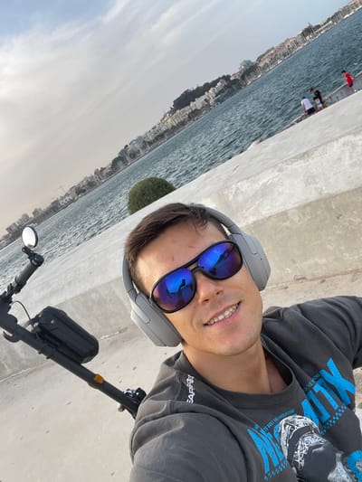 A man wearing headphones and sunglasses takes a selfie on a sunny day. He is seated on an electric scooter near a body of water, possibly the Mediterranean Sea, with a city skyline in the background. The city appears to be Cartagena, Spain, based on the architectural style and port activity visible. The man's reflection is visible in his sunglasses. He is smiling and wearing a dark gray t-shirt. In the background, several people are visible near the water's edge. The environment is bright and appears to be a promenade or walkway.