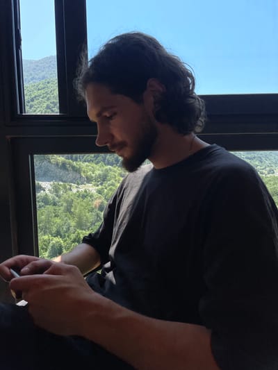 A young man with dark, medium-length curly hair and a beard is sitting indoors, looking down at his hands which are holding a white object, possibly a cigarette or vape pen. He is wearing a dark t-shirt and a silver chain around his neck. He is positioned in front of a large window with a view of a lush, green mountainous landscape under a clear blue sky. The window frame is black. The location is Meneso, Georgia.