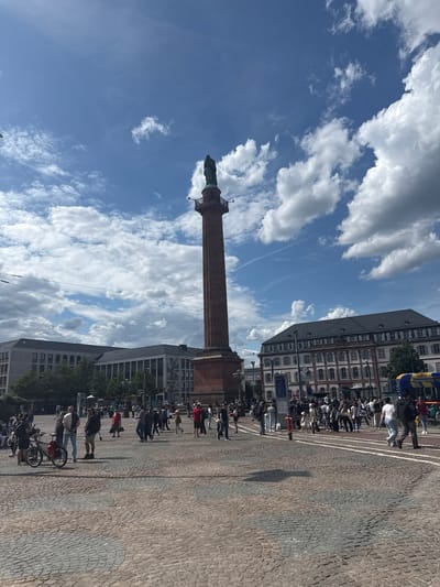 This image captures a vibrant daytime scene on Luisenplatz in Darmstadt, Germany. The central focal point is the towering, reddish-brown Ludwigsmonument (Langer Ludwig) column, crowned by a green-patinaed statue, likely depicting Grand Duke Ludwig I. A small, dark-railed observation platform is visible just beneath the statue.

The expansive foreground and midground consist of a cobblestone-paved square, exhibiting variations in color and texture, with faint lighter lines or patterns on its surface. A significant number of pedestrians, dressed in casual attire, are distributed across the square, walking in various directions, standing, and interacting. On the left, a person is standing near a black bicycle equipped with a red bag on its rear rack.

To the left of the monument, several modern, multi-story buildings with light-toned facades, dark roofs, and numerous windows line the square. One of these buildings features a large, dark mural or graphic on its side. To the right of the monument stands a classical-style, multi-story building characterized by a prominent dark roof, a blend of white and reddish-brown brickwork, and many windows fitted with white shutters. A segment of a blue and yellow public transport vehicle, possibly a bus or tram, is visible on the far right.

Throughout the scene, street furniture is present, including lampposts, information boards, and a red fire hydrant. Multiple black overhead wires, indicative of tram or trolleybus lines, traverse the upper portion of the image. The sky is clear and blue, interspersed with fluffy white cumulus clouds, suggesting bright, sunny weather. The strong sunlight casts visible shadows from people and structures onto the square. Green foliage and trees are visible beyond some of the buildings.