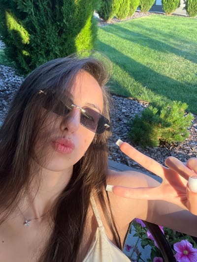 In Novooleksandrivka, Ukraine, a young woman with long, dark hair and sunglasses is taking a selfie outdoors. She is wearing a light-colored top and a delicate silver necklace. Her lips are pursed, and she is making a peace sign with her right hand, which has long, white-painted nails. The background features a manicured lawn, trimmed evergreen bushes, and shadows cast by sunlight, suggesting it is daytime.