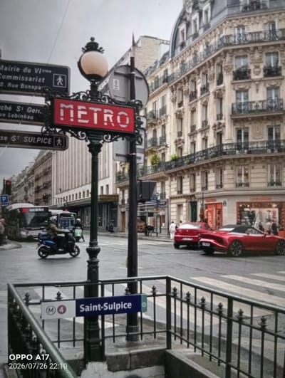 The image depicts a street scene in Paris, France, not Құмөзек, Kazakhstan. In the foreground, a Metro station sign for "Saint-Sulpice" is visible, marked with "M 4". A black metal railing surrounds the entrance to the station. Further back, a street runs horizontally with two red cars, one a convertible, parked or driving on it. A bus is visible on the left side of the street, along with a person riding a scooter. In the background, the architecture of Parisian buildings is prominent, with ornate facades and balconies. The sky is overcast. Signs in French indicate directions to the "Mairie du VIème", "Commissariat", and "Marché St. Ger. rue Lobineau". The scene captures an urban environment with typical city traffic and infrastructure.
