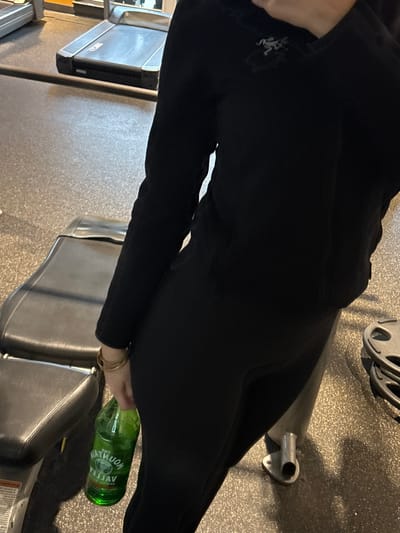 The image depicts a person wearing black leggings with a logo on the thigh. They are seated on a black weight bench at a gym. A green glass water bottle rests on the bench near their hand, adorned with gold bracelets. A treadmill is visible in the background. Other gym equipment is also visible, including what appears to be a weight rack and various gym machines. The floor appears to be gray.