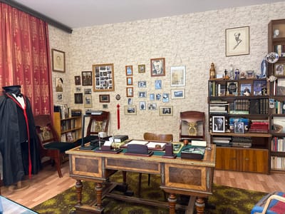 The image shows a meticulously arranged room, likely a study or office, within a museum setting in Ovsianka, Russia, specifically the former residence of Viktor Astafyev. The room is filled with personal effects and memorabilia, creating a strong sense of the writer's presence.
Dominating the scene is a large, ornate wooden desk with intricate carvings and inlays. On the desk, a green blotter holds several items: a pen set, a notepad, a paperweight, and a small wooden object. To the right of the desk, a metal lamp with a black shade is positioned.
Behind the desk, two antique wooden chairs with gold ornate detailing are placed. One chair is a high-backed armchair with dark green upholstery, while the other appears to be a more upright chair.
The walls are adorned with numerous framed photographs and portraits, mostly black and white, depicting individuals and groups, likely family members and notable figures. A large, dark red curtain with gold patterns hangs on the left side of the room, framing a mannequin dressed in a black academic robe with red trim and a graduation cap.
A tall bookshelf to the right is filled with books, many of which appear to be classic literature and historical texts. Decorative objects are interspersed among the books, including figurines, a blue and white decorative plate with a Russian emblem, and a stylized sculpture of a fish.
The floor is covered with a patterned green and gold rug, and the surrounding walls are wallpapered with a light, textured pattern. The overall impression is one of a preserved historical space, offering a glimpse into the life and work of a respected Russian writer.