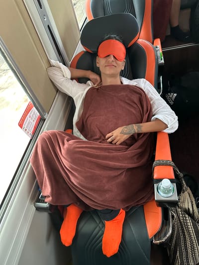 A person wearing an orange eye mask and orange socks is reclining in a seat on a train. They are covered with a maroon blanket and wearing a white shirt. The seat is black with orange accents. A bottle of water is in a cup holder attached to the seat. A safety and emergency instruction card is visible on the window. The train appears to be in motion.