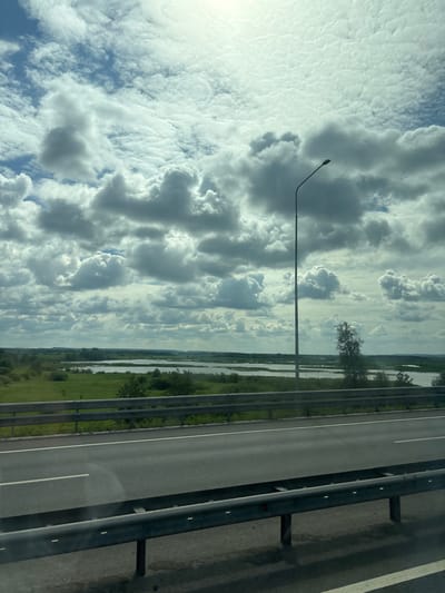 The image captures a view from a highway looking towards a vast landscape under a dramatic, cloud-filled sky. The highway itself is visible in the foreground, featuring asphalt with white lane markings and a metal guardrail. Beyond the highway lies a green grassy field dotted with scattered trees and bushes. Further in the distance, a body of water, likely a river or lake, stretches across the horizon, reflecting the sky. The sky is dominated by a complex pattern of cumulus clouds, with patches of blue sky peeking through. A tall, slender lamppost stands on the right side of the highway, its light fixture pointing upwards. The scene is devoid of any people or discernible activity, presenting a serene, natural environment. It is difficult to definitively place this specific location within Menzelinsk, Russia, without further context or identifiable landmarks.