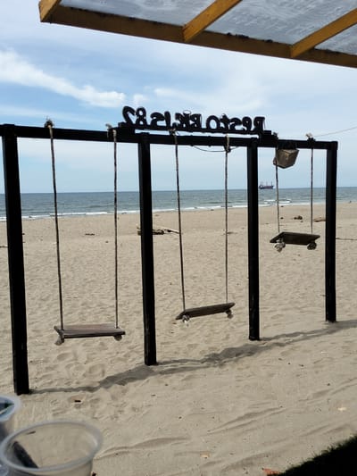 A wooden swing set with three seats stands on a sandy beach, facing the ocean. The sky is blue with scattered white clouds. The sea is calm, with a few small waves. A large ship is visible in the distance. The swing set is made of black metal posts and crossbars. The seats are wooden planks suspended by ropes. A small wooden box is hanging from the top bar of the swing set. In the foreground, two plastic cups are visible on the sand. The scene appears to be a quiet, relaxed setting, possibly a beachside restaurant or recreational area.
