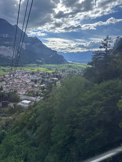 The image is an aerial view looking down on the town of Meiringen, Switzerland. The perspective suggests the photo was taken from a cable car or gondola, with thick black cables visible in the foreground of the frame, stretching diagonally from the top left.
The town is nestled in a valley, characterized by a dense collection of buildings with red-tiled roofs and lighter-colored walls, typical of Swiss architecture. Some industrial buildings with flat roofs and a train station are also discernible within the urban area. Beyond the town, extensive green fields stretch out, bordered by the steep, tree-covered slopes of mountains. In the distance, a range of hazy, blue mountains forms the horizon, under a dramatic sky filled with a mix of dark and bright clouds, with patches of sunlight breaking through. The overall environment is mountainous and rural, with a clear contrast between the developed town and the natural landscape. There are no people visible in the frame.