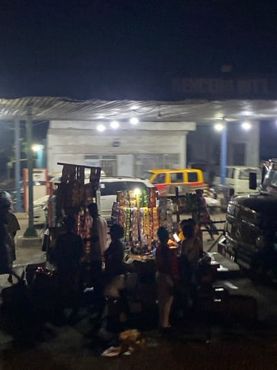 A night street scene shows a building illuminated by several lights, next to what appears to be a vendor stall overflowing with colorful packaged goods. People are gathered around the stall, possibly shopping. Parked vehicles are also visible, including a yellow and red vehicle and others behind the vendor. The overall scene is somewhat dark and blurry.