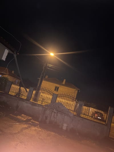 The image captures a nighttime scene in Pishtigovo, Bulgaria, featuring a street illuminated by a single, bright street lamp. The street lamp, emitting a strong orange-yellow glow and significant lens flares, is positioned on a utility pole towards the left of the frame. Numerous power lines extend from the pole, crisscrossing the dark sky.
To the right, a multi-story building, likely a residence, stands under the artificial light. Its facade appears to be constructed of yellowish-tan brick or stone, with visible windows and decorative wrought-iron railings on what seem to be balconies or a fence line. These dark, ornate metal structures add a decorative element to the property.
Behind the fence, a dark-colored car is partially visible, parked in what appears to be a driveway or yard. Its rear lights and a faint license plate are discernible, with the plate possibly indicating a Bulgarian registration (e.g., beginning with 'CA' or 'CB' for Sofia or Vidin). Some dark vegetation can be seen behind the car. Various rough-textured wall surfaces, possibly concrete or stucco, form parts of the property boundaries and lower structures.
The overall atmosphere is dark, with deep shadows contrasted by the intense light from the street lamp. Some surfaces appear slightly damp or reflective, suggesting recent precipitation or moisture in the air. There are no people visible, and no specific activity is taking place; the image presents a static street view after dark.